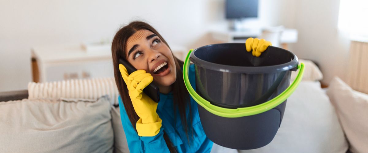 Shocked woman calling plumber while collecting water leaking from ceiling using utensil (2)